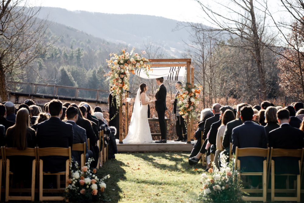 Guests seated in wooden chairs watch a couple exchange vows beneath a floral-adorned chuppah with sweeping Catskills mountain views, the kind of ceremony moment that comes together when planning a wedding weekend in Hudson Valley.
