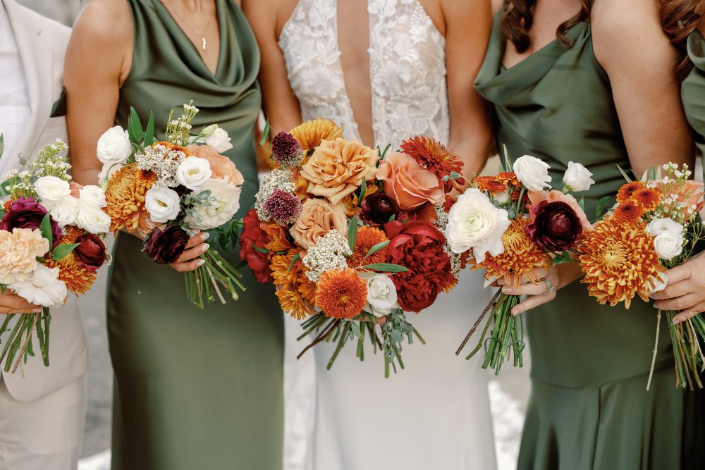 Bride and bridesmaids in sage green dresses hold vibrant fall bouquets with orange, burgundy, peach, and white blooms, a warm color palette perfect for a Hudson Valley wedding weekend in autumn.