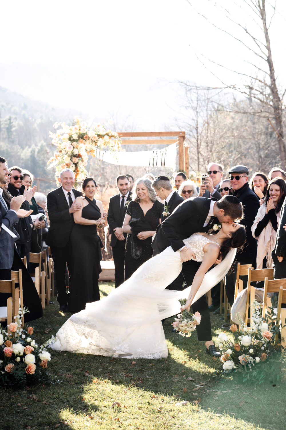 Groom dips bride for a kiss as guests cheer and applaud at the end of their outdoor Hudson Valley wedding ceremony, with a floral chuppah and mountain scenery in the background.
