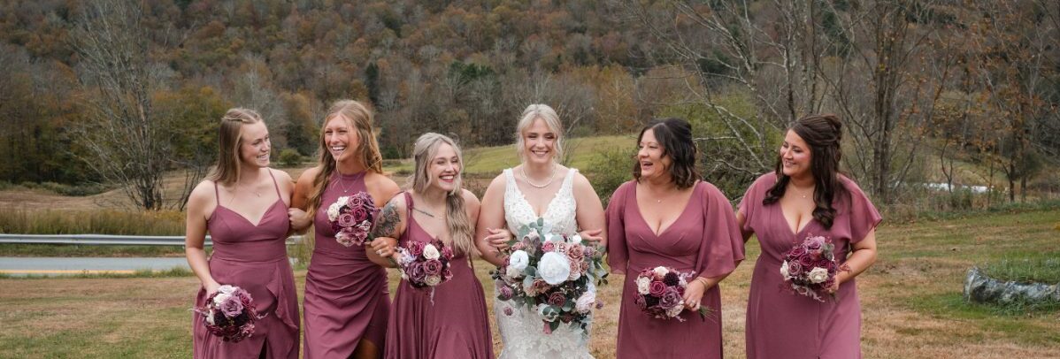 Bride and bridesmaids walk together across an open field in matching mauve dresses, holding bouquets and smiling. A relaxed portrait moment that fits naturally into the pacing of a well-organized wedding weekend itinerary.