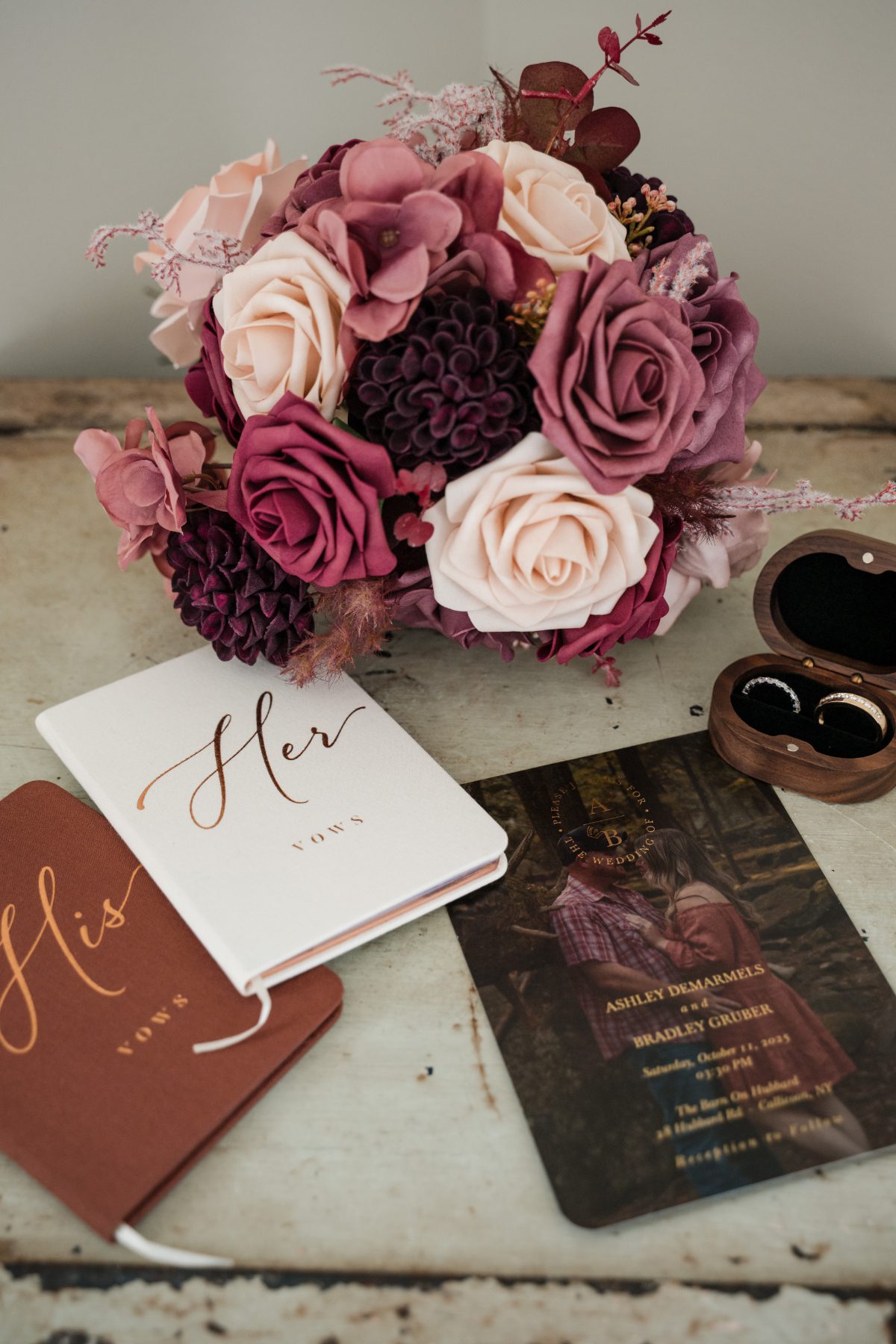 Close-up of a bridal bouquet with mauve, blush, and deep plum florals beside vow books, rings, and an invitation suite. Soft textures and warm tones reflect thoughtful details often planned into a wedding weekend itinerary.