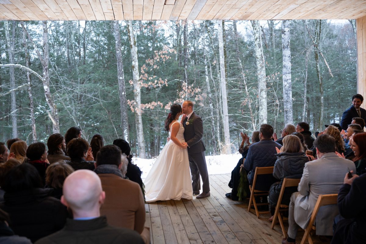 Couple shares a kiss during an indoor ceremony overlooking a wooded landscape, surrounded by seated guests. The setting feels calm and intimate, a moment that fits seamlessly into a well-planned wedding weekend itinerary.