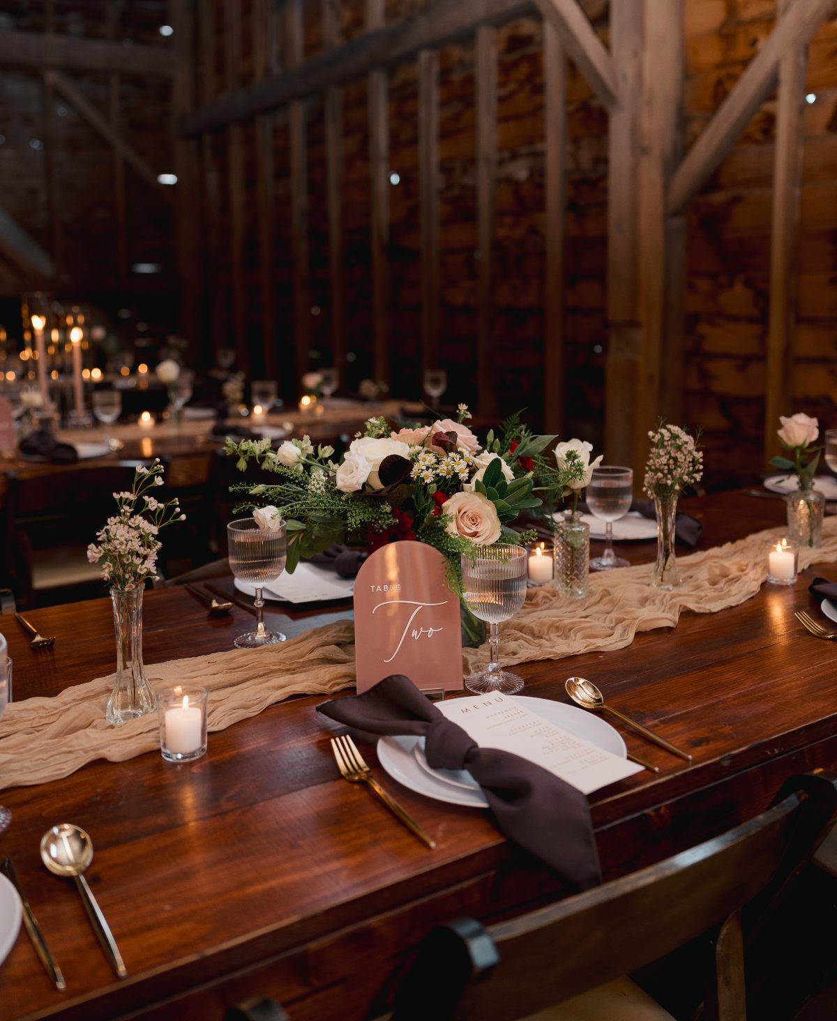 Reception table styled with candles, florals, and neutral linens inside a rustic wood barn. The setup feels warm and intentional.