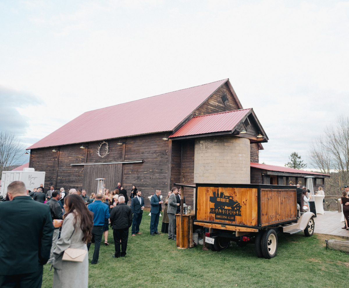 Guests mingle outside near The Barn on Hubbard barn venue with a mobile bar, holding drinks and chatting before the reception. A casual, social moment that highlights the flow and guest experience of a well-structured wedding weekend itinerary.