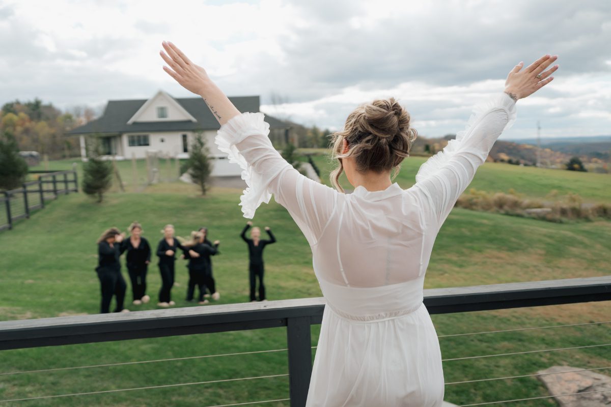 Bride stands on a balcony with arms raised, overlooking a scenic landscape as her bridal party gathers below. A quiet, in-between moment that reflects the breathing room built into a balanced wedding weekend itinerary.