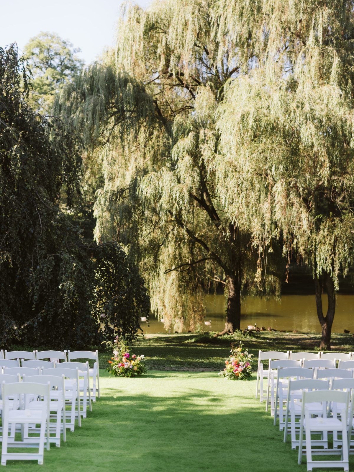 Outdoor ceremony setup at Buttermilk Falls with white chairs and floral arrangements by a pond and willow trees, ideal for destination weddings in Hudson Valley.
