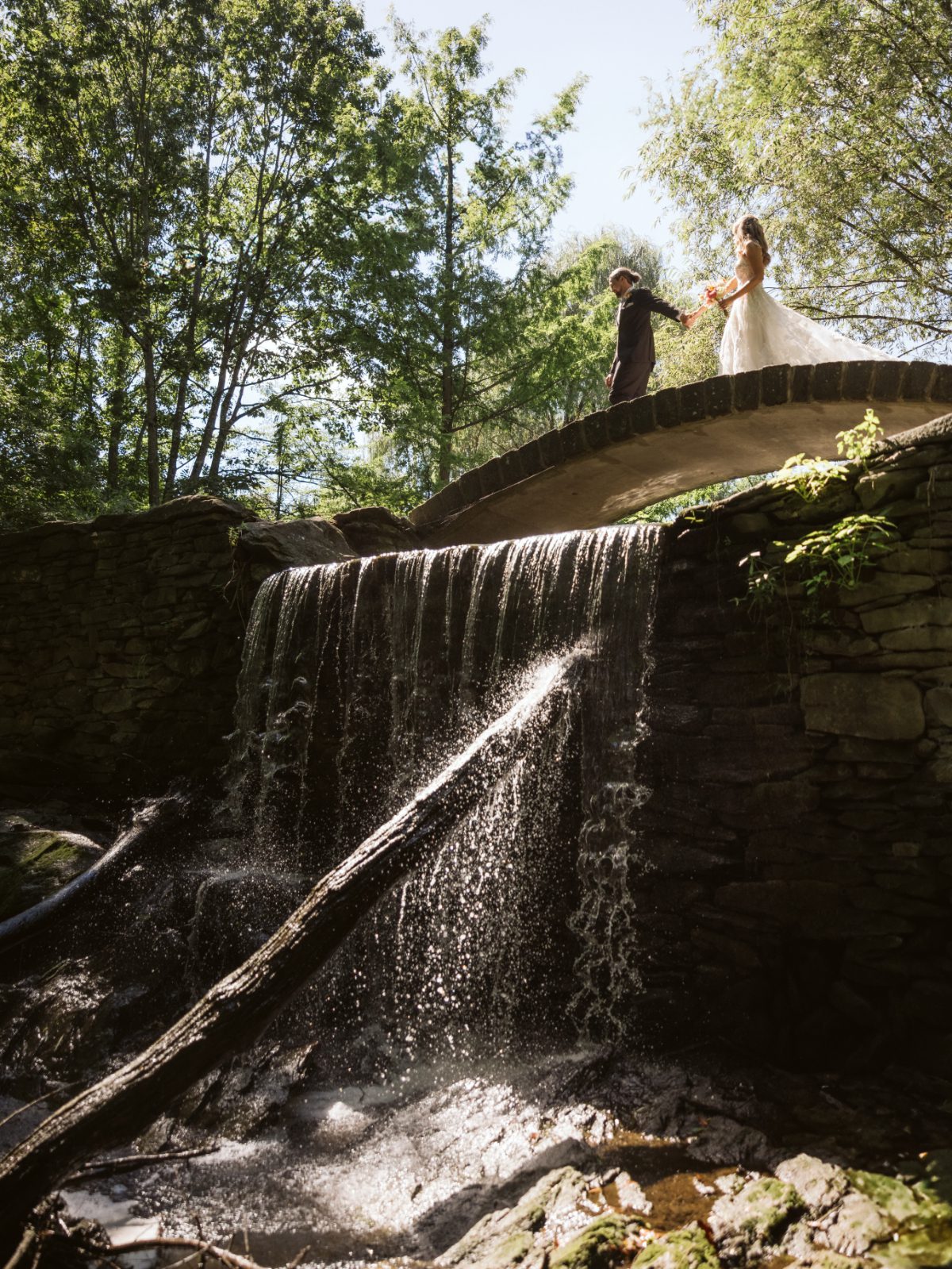 Couple walks across a stone bridge above a waterfall at Buttermilk Falls, surrounded by trees and sunlight, creating a romantic setting for destination weddings in Hudson Valley.