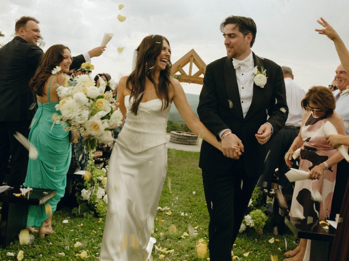 Bride and groom walk down the aisle smiling as guests toss flower petals during an outdoor ceremony, capturing the joy and energy often seen at destination weddings in Hudson Valley.