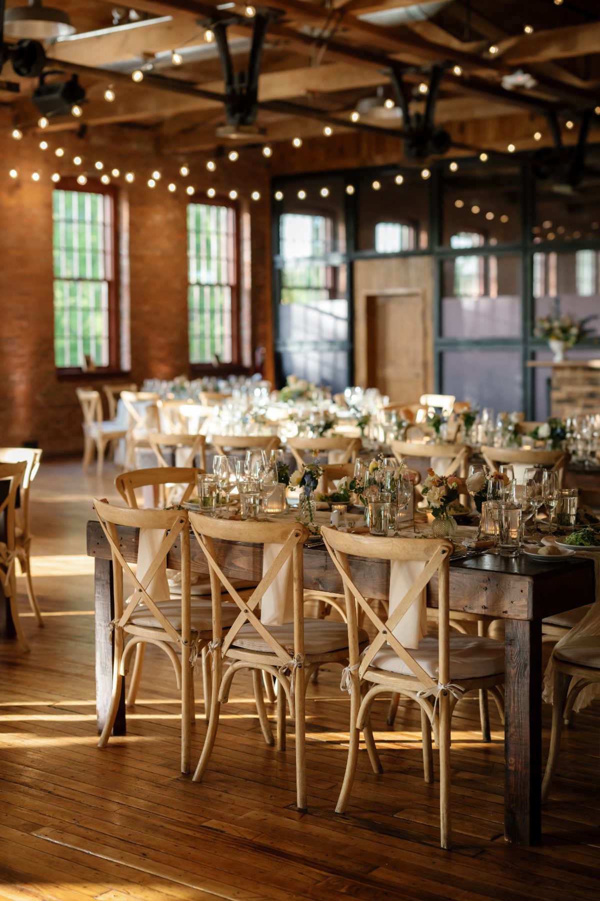 Warm, light-filled reception space with wood tables, cross-back chairs, and string lights in a brick venue, creating an elegant setting often seen in destination weddings in Hudson Valley.