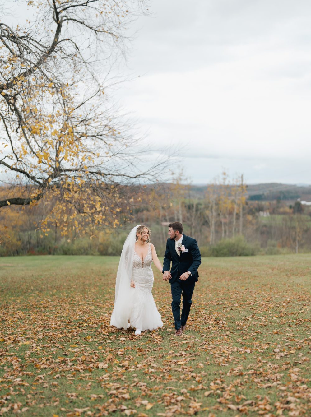 A bride and groom walk hand in hand across a leaf‑covered field surrounded by bare trees and distant hills. 