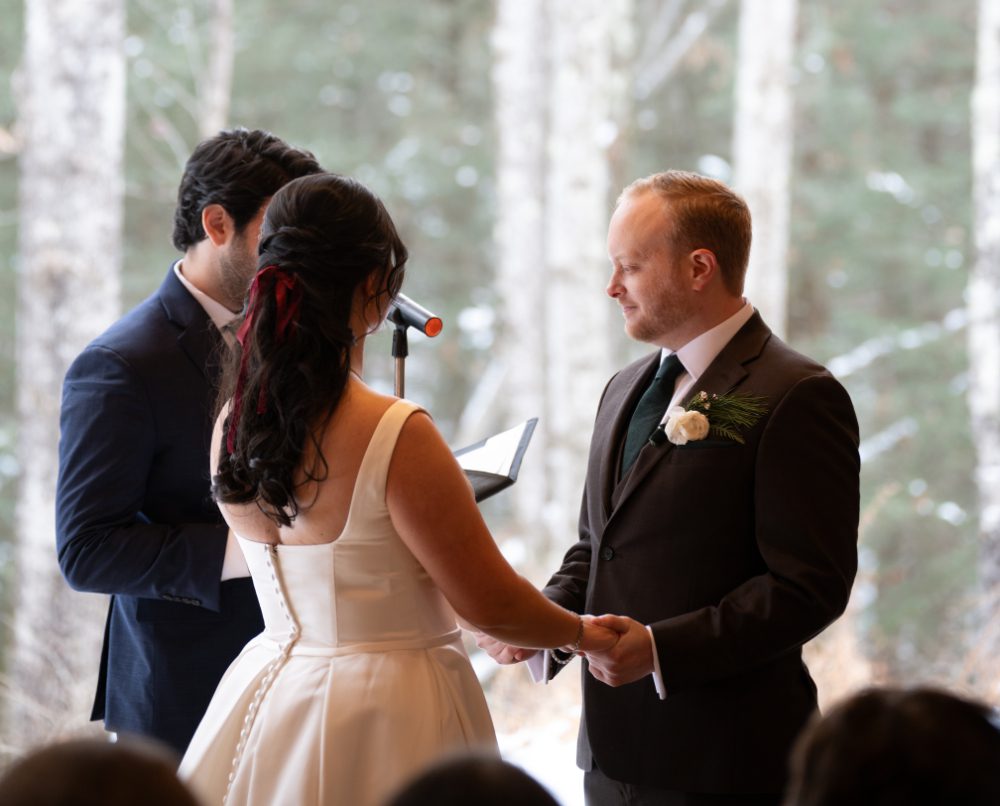 The bride and groom hold hands and exchange vows during their Audrey's Farmhouse wedding, with a woodland backdrop and the officiant standing beside them.