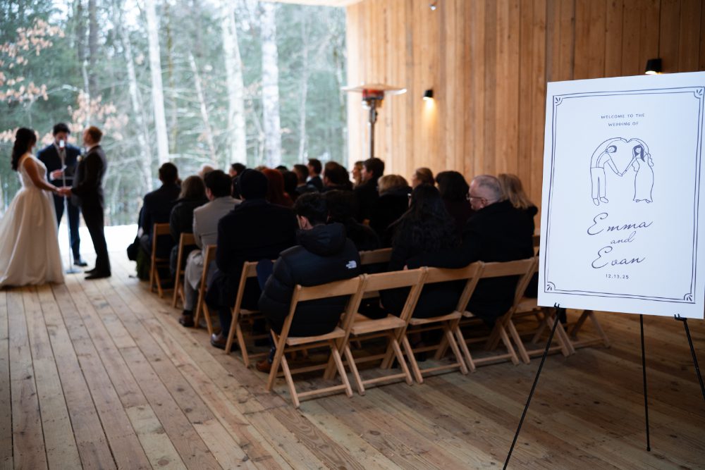 Guests watch the bride and groom exchange vows inside the wooden ceremony pavilion, with a custom illustrated wedding welcome sign displayed beside the seating.
