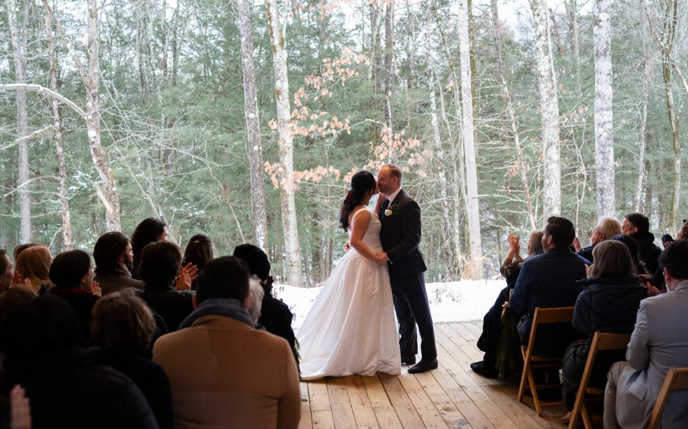 The bride and groom share their first kiss as wedding guests applaud inside the open-air wooden pavilion at their Audrey's Farmhouse wedding.
