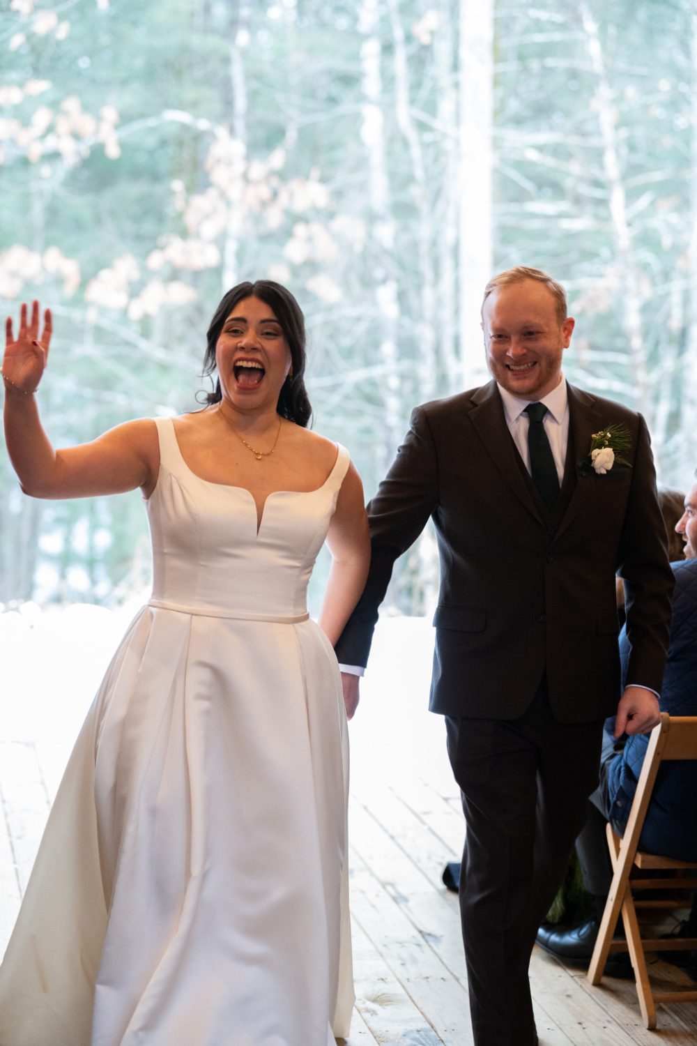 The bride waves excitedly while the groom smiles as they walk hand in hand down the aisle after their winter wedding ceremony in the Hudson Valley.