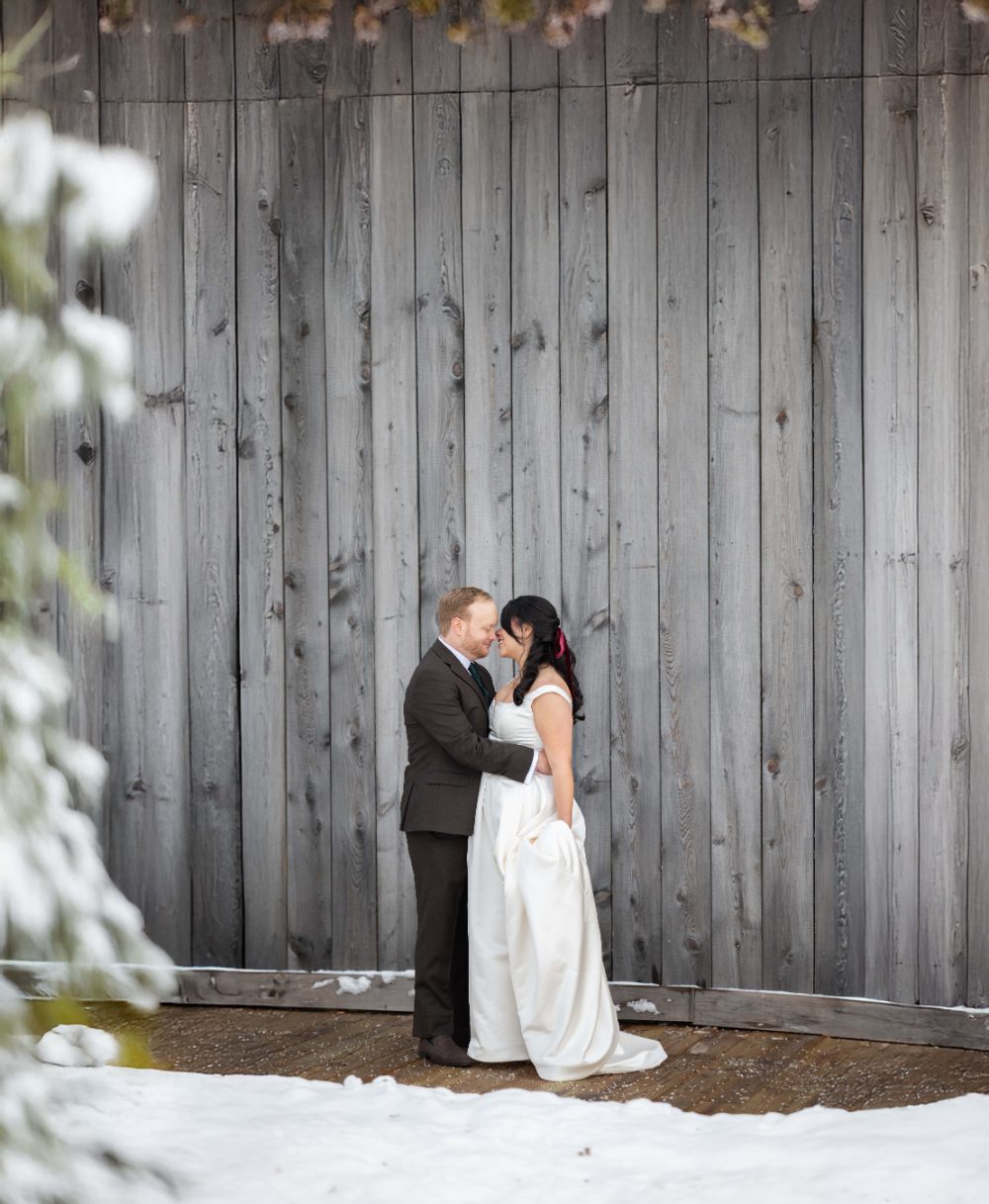 Bride and groom embrace in front of a rustic gray wooden wall during their Audrey's Farmhouse wedding, with snow along the deck and bride holding her flowing gown.