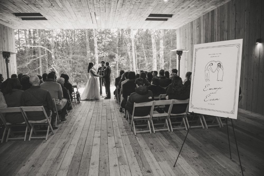 Black-and-white wide-angle view of an Audrey's Farmhouse wedding ceremony, featuring the bride and groom exchanging vows, seated guests, and a custom welcome sign.