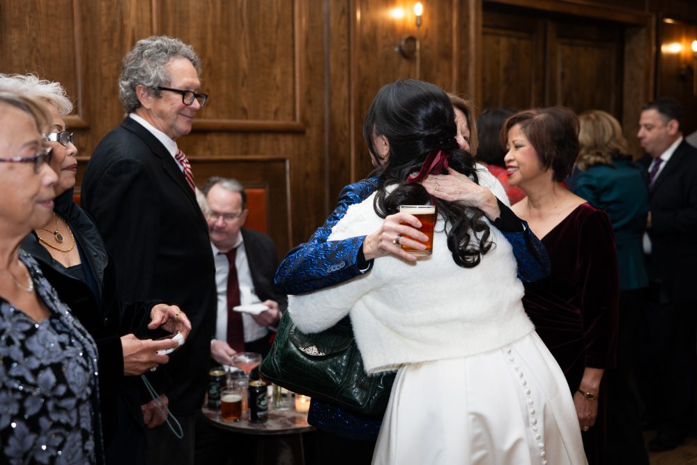 The bride embraces a wedding guest during cocktail hour at Friends and Neighbors, surrounded by family and friends in a warmly lit, wood-paneled room.