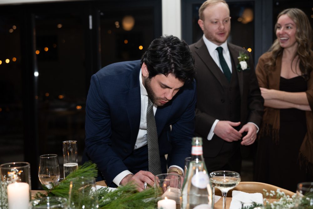 A wedding guest signs a card at a reception table while the groom and another guest look on, surrounded by winter greenery and candlelight.