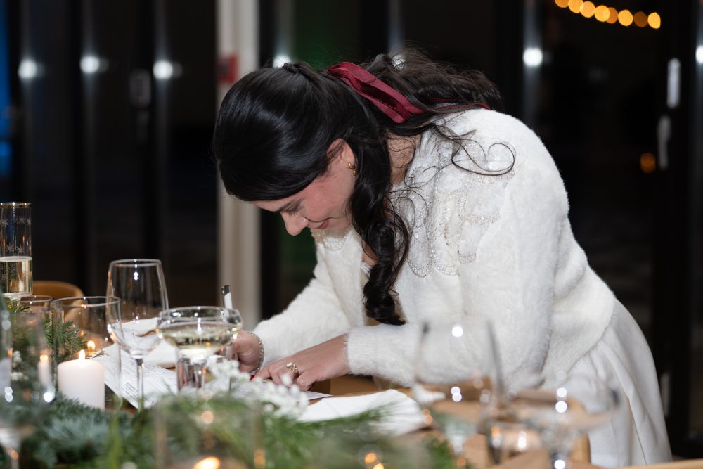 The bride, wearing a white wedding gown and embroidered cardigan, writes a note at a candlelit table during her Audrey's Farmhouse wedding reception.
