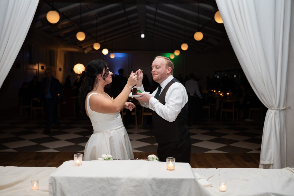 The bride playfully feeds the groom wedding cake during their Audrey's Farmhouse reception, surrounded by white drapery, warm pendant lights, and a checkered dance floor.