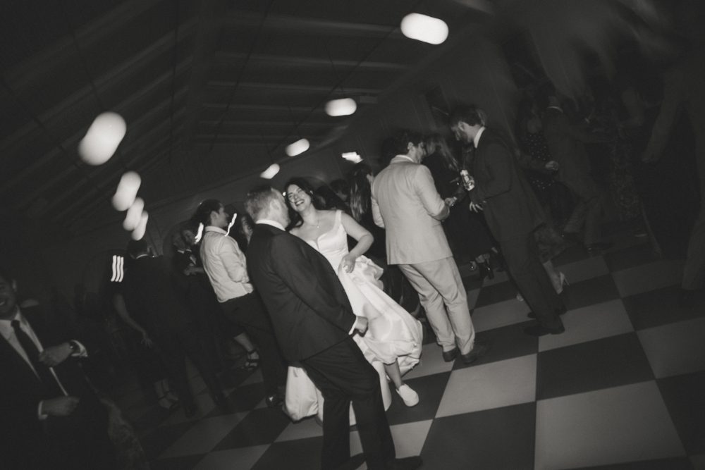 Black-and-white photograph of bride and groom laughing and dancing with guests on the checkered floor at their wedding reception.