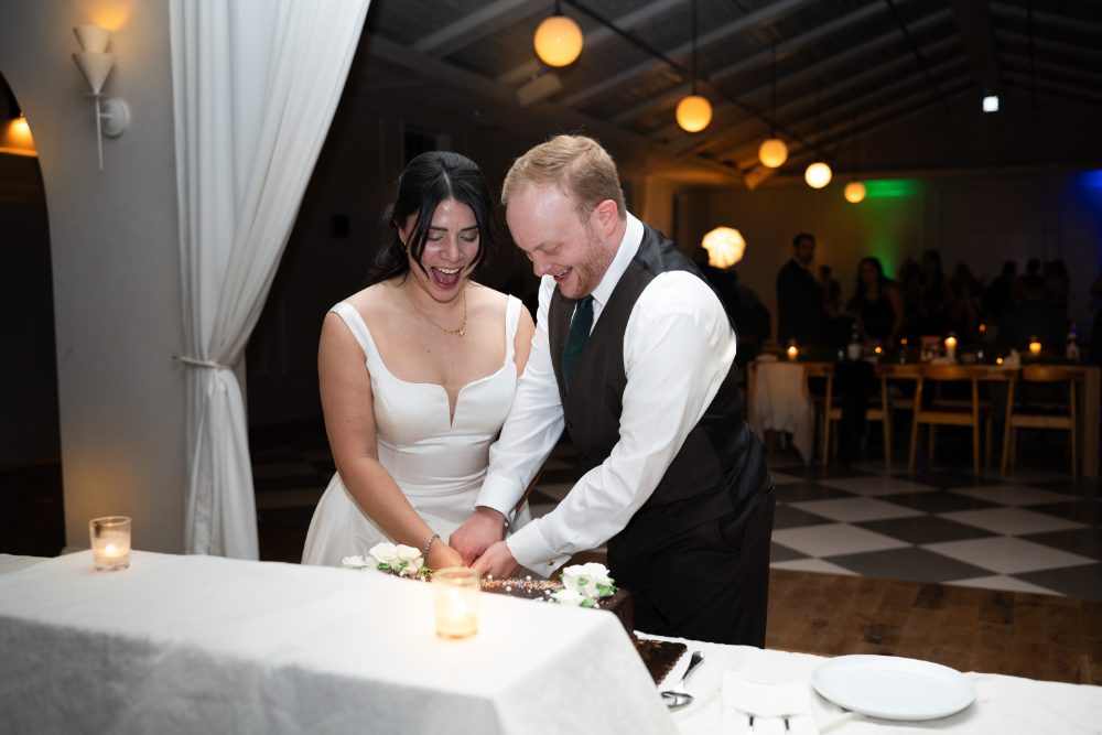 The bride and groom laugh as they cut their chocolate wedding cake together during their Audrey's Farmhouse wedding reception.