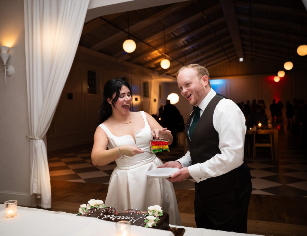 The bride laughs while serving the groom a slice of colorful layered wedding cake during their reception at Audrey's Farmhouse in the Hudson Valley.