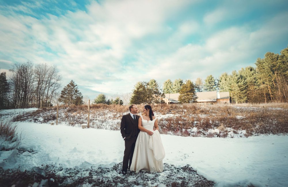 Bride and groom stand together in a snow-covered field during their Audrey's Farmhouse wedding in Stone Ridge, New York, surrounded by winter trees beneath a cloudy blue sky.