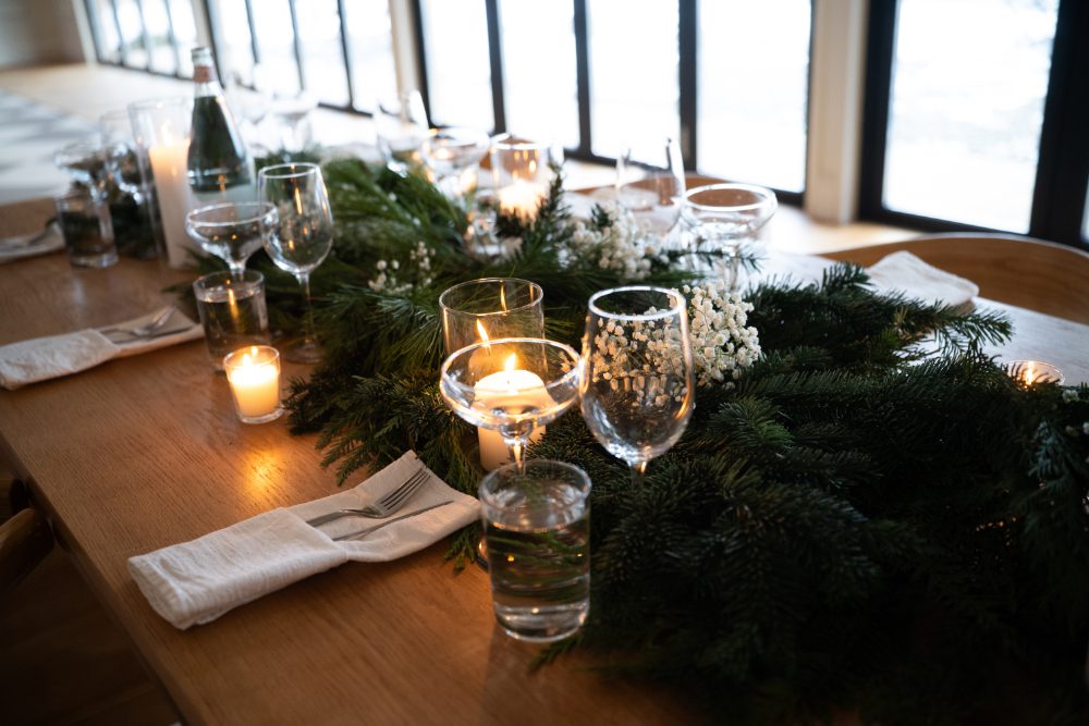 Glowing pillar candle surrounded by fresh winter greenery and white flowers at an Audrey’s Farmhouse wedding reception.