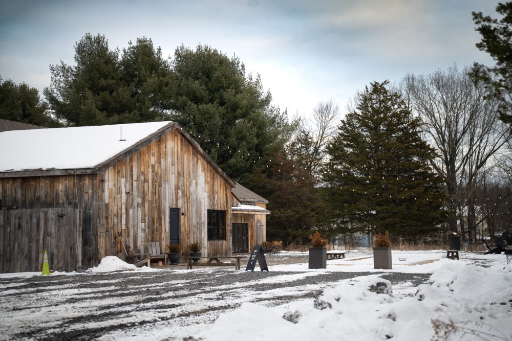 A rustic wooden event barn at Audrey's Farmhouse in the Hudson Valley, surrounded by snow-covered grounds, evergreen trees, and string lights on a quiet winter wedding day.