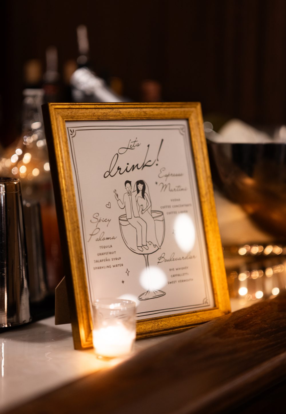 A gold-framed signature cocktail menu featuring illustrated bride and groom artwork and personalized drinks sits beside a candle at an Audrey's Farmhouse wedding.