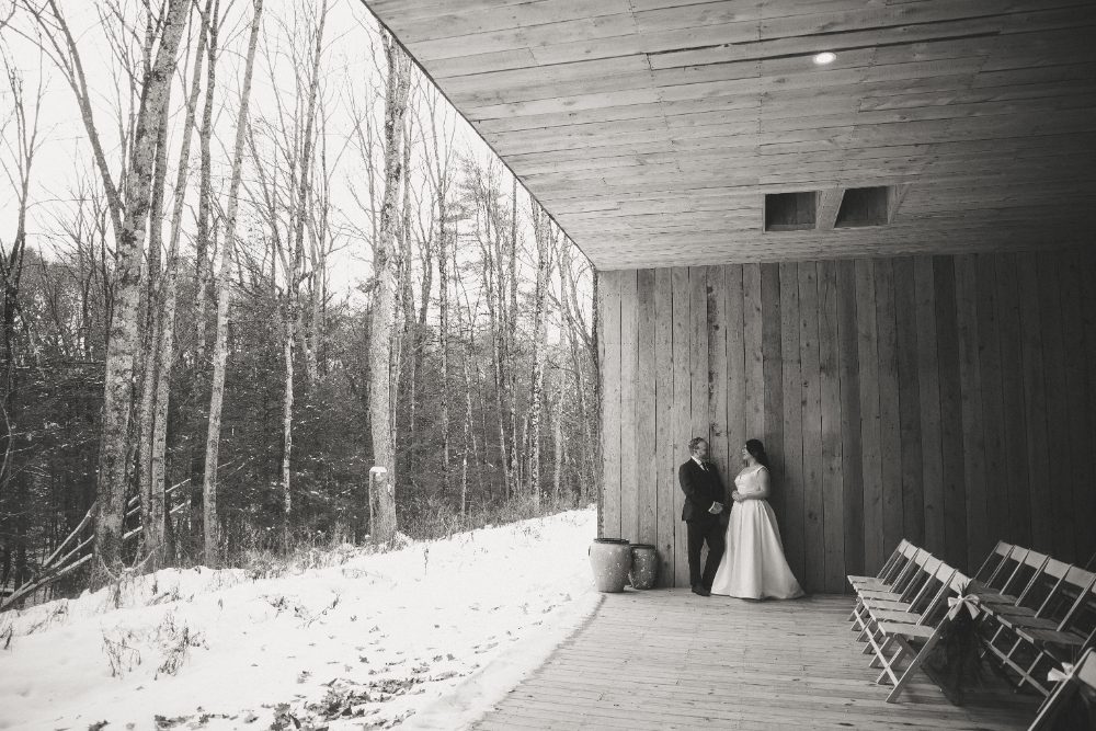 Black-and-white wedding portrait of bride and groom standing beneath a wooden pavilion at Friends and Neighbors, with snow-covered ground and bare winter trees nearby.