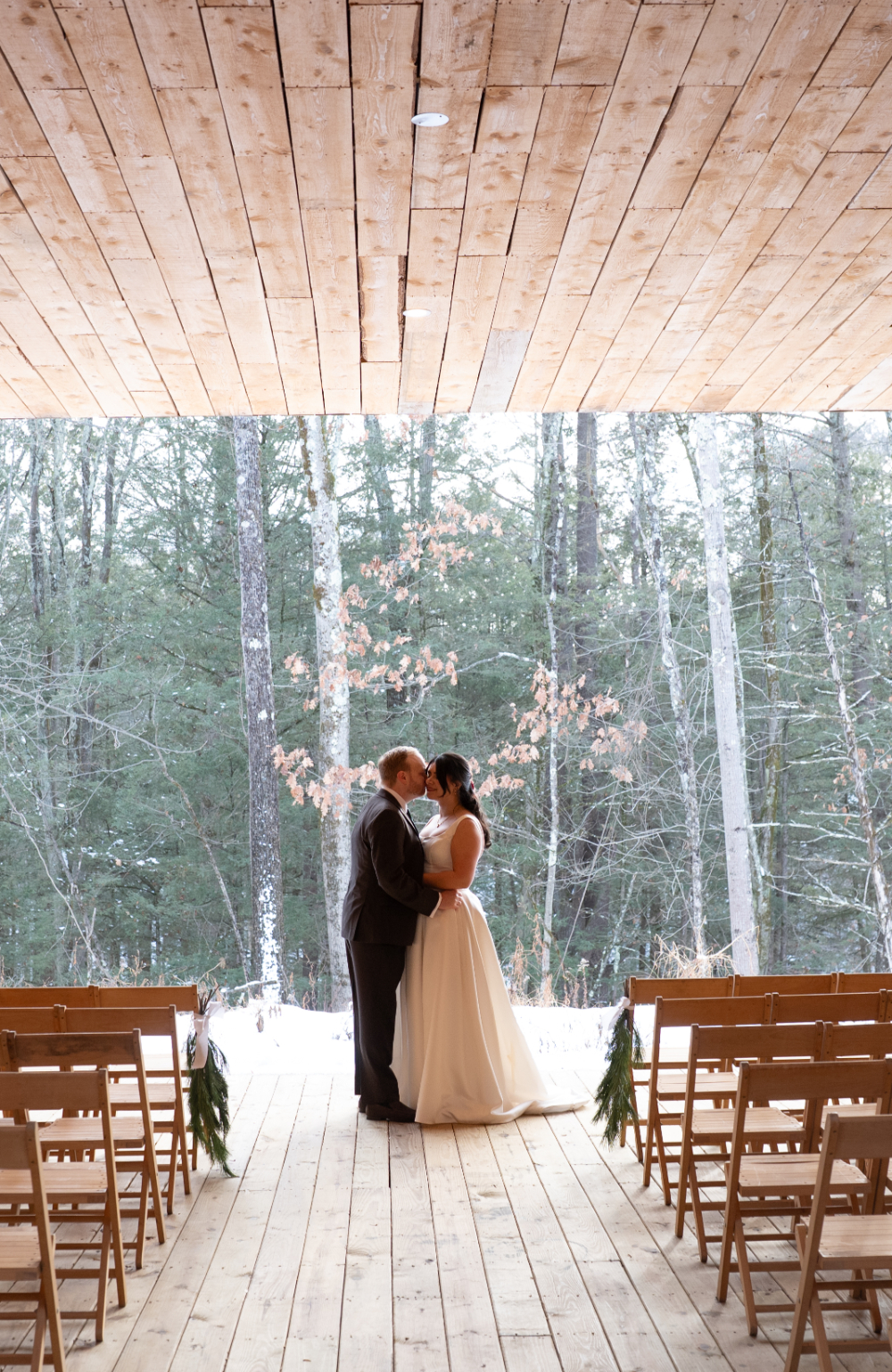 Bride and groom stand together during their Audrey's Farmhouse wedding ceremony in a wooden pavilion overlooking the winter forest, with greenery lining the aisle.